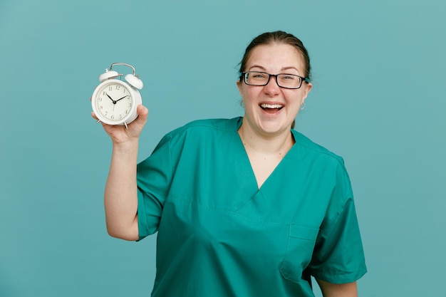 Молодая женщина-медсестра в медицинской форме со стетоскопом на шее, держащая будильник, смотрит в камеру, счастливая и взволнованная, смеясь, стоя на синем фоне