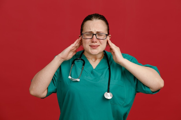 Young woman nurse in medical uniform wearing glasses with stethoscope around neck looking annoyed closing ears with hands standing over red background