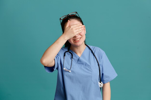 Young woman nurse in medical uniform with stethoscope around neck covering eyes with hand smiling cheerfully standing over blue background