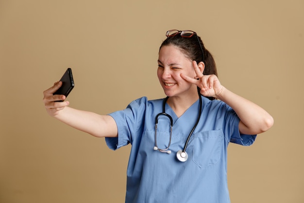 Young woman nurse in medical uniform with stethoscope around neck doing selfie using smartphone smiling cheerfully showing vsign standing over brown background