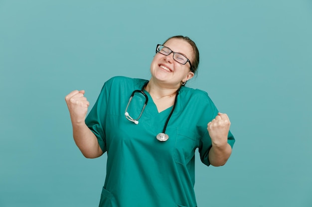 Young woman nurse in medical uniform with stethoscope around neck happy and excited clenching fists rejoicing her success standing over blue background