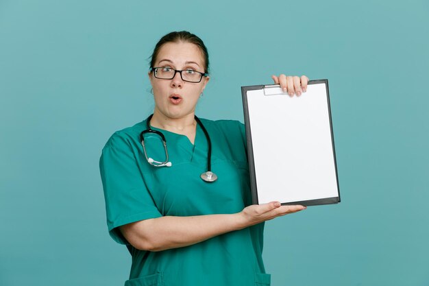 Giovane donna infermiera in uniforme medica con stetoscopio intorno al collo che tiene appunti guardando la fotocamera sorpresa in piedi su sfondo blu