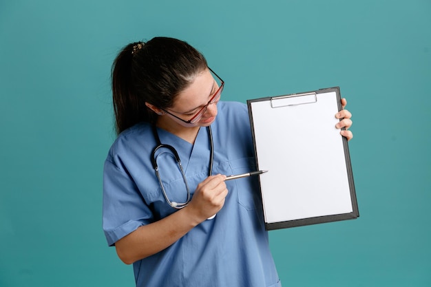 Young woman nurse in medical uniform with stethoscope around neck holding clipboard with blank page and pen looking at clipboard with interest standing over blue background