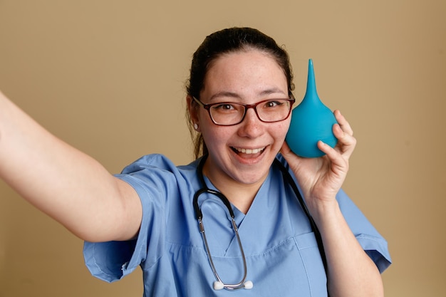 Young woman nurse in medical uniform with stethoscope around neck holding enema looking at camera happy and cheerful smiling standing over brown background