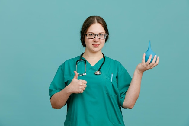 Young woman nurse in medical uniform with stethoscope around neck holding enema looking at camera smiling confident showing thumb up standing over blue background