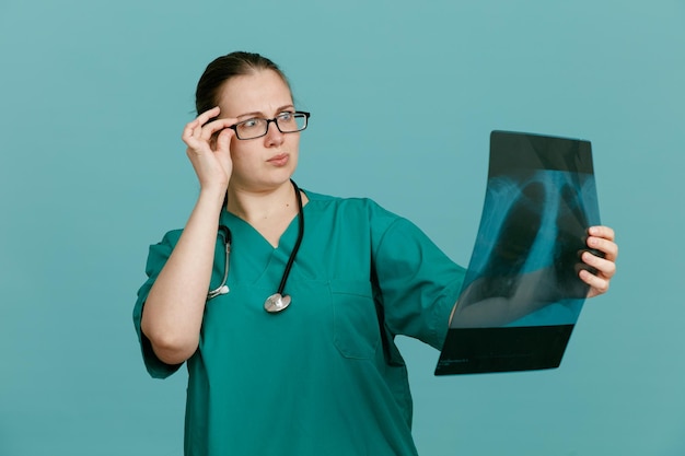 首の周りに聴診器を持った医療制服を着た若い女性看護師が、青い背景の上に立って混乱して心配している肺のレントゲン写真を持っています