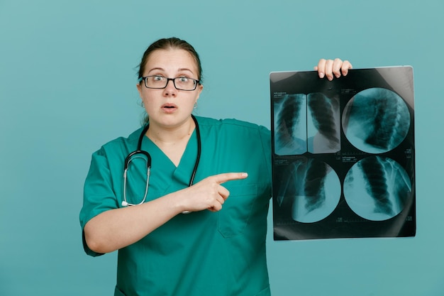 Молодая женщина-медсестра в медицинской форме со стетоскопом на шее держит рентген легких, указывая указательным пальцем на него, удивленная и обеспокоенная стоя на синем фоне