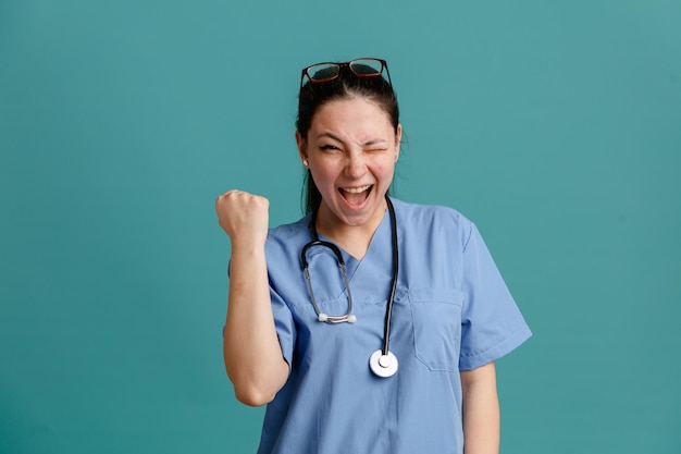 Young woman nurse in medical uniform with stethoscope around neck looking at camera happy and excited clenching fist rejoicing her success standing over blue background