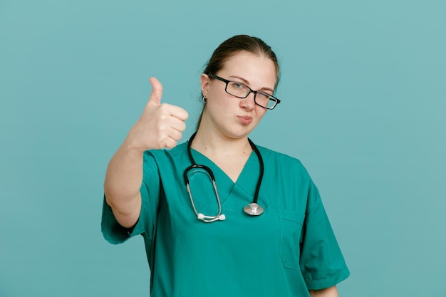 Young woman nurse in medical uniform with stethoscope around neck looking at camera happy and positive smiling showing thumb up standing over blue background