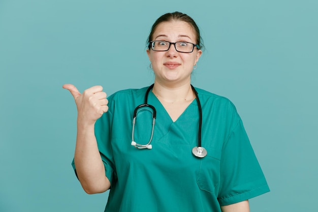 Young woman nurse in medical uniform with stethoscope around neck looking at camera happy and surprised pointing with thumb to the side standing over blue background