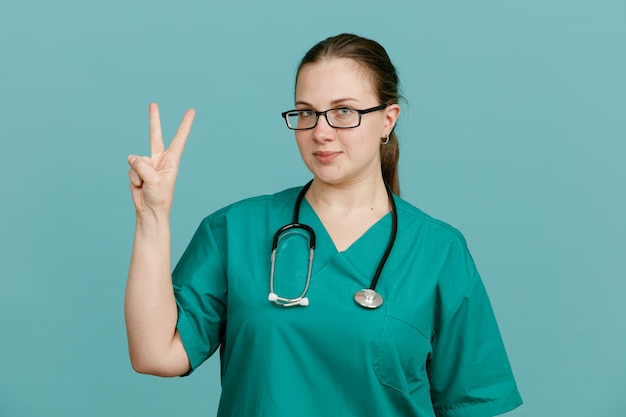 Young woman nurse in medical uniform with stethoscope around neck looking at camera smiling confident showing number two or vsign standing over blue background