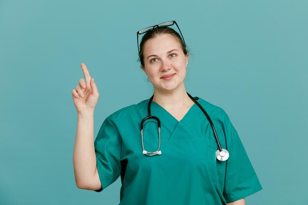 Giovane donna infermiera in uniforme medica con uno stetoscopio intorno al collo che sembra sorridente fiducioso che mostra il dito indice in piedi su sfondo blu