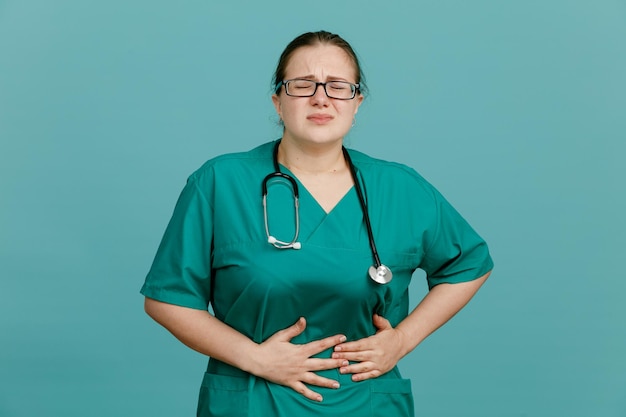 Young woman nurse in medical uniform with stethoscope around neck looking unwell touching her belly feeling pain standing over blue background