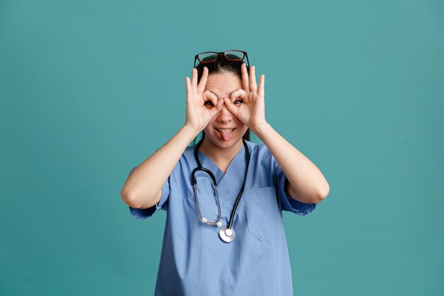 Young woman nurse in medical uniform with stethoscope around neck making binocular gesture sticking out tongue looking through fingers standing over blue background