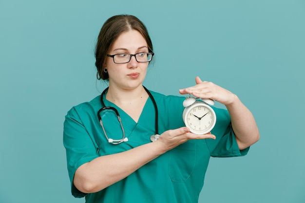 Giovane donna infermiera in uniforme medica con stetoscopio intorno al collo che mostra sveglia guardandolo confuso in piedi su sfondo blu