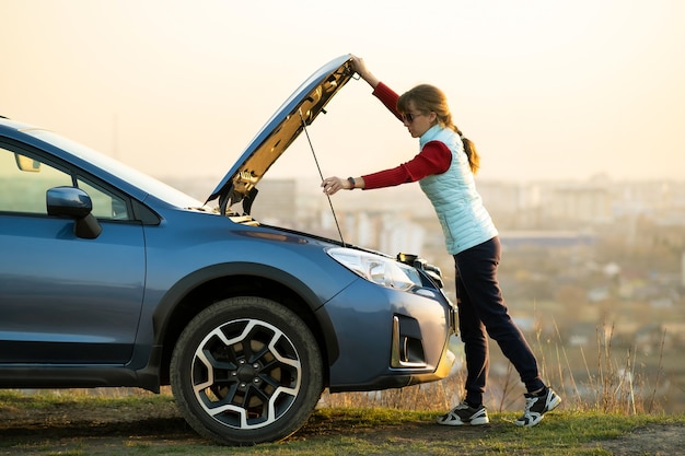 Молодая женщина открывает капот сломанной машины, имея проблемы с ее транспортным средством. Женщина-водитель, стоящая возле авто с поднятым капюшоном.
