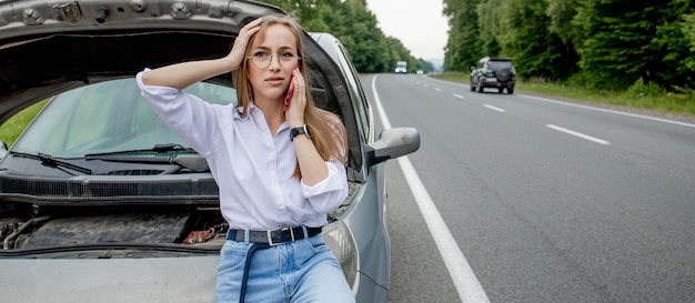 壊れた車のボンネットを開ける若い女性が自分の車に問題を抱えています。壊れた車の近くで電話で話している心配の女性。壊れた車の近くに立っている女の子モデルが自動車サービスを求めています。