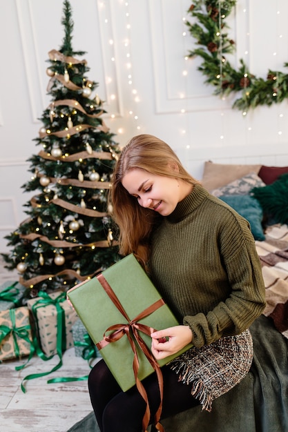 若い女性が新年の雰囲気の中でクリスマスプレゼントを開き、喜ぶ。女性は明るく居心地の良い寝室にいて、クリスマスのために美しく装飾されています。