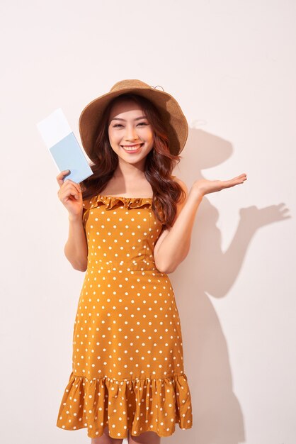 A young woman in a orange polka dots dress with a straw hat on her head is holding a passport and plane tickets