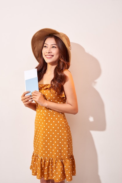 A young woman in a orange polka dots dress with a straw hat on her head is holding a passport and plane tickets