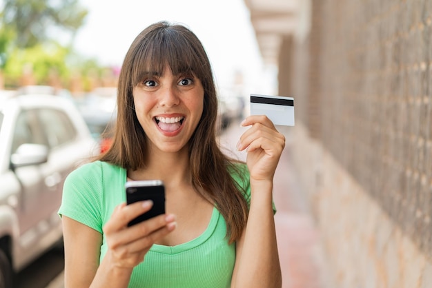 Young woman at outdoors buying with the mobile and holding a credit card with surprised expression