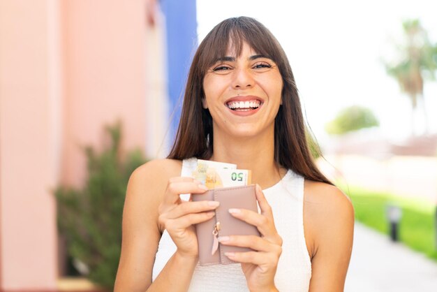 Молодая женщина на улице держит бумажник с деньгами со счастливым выражением лица