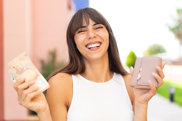 Молодая женщина на улице держит бумажник с деньгами со счастливым выражением лица