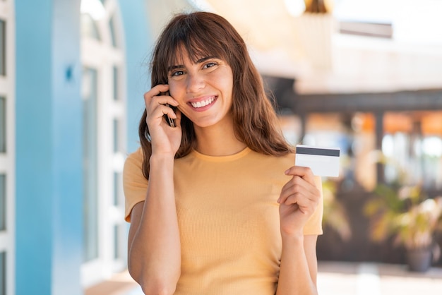 Young woman at outdoors keeping a conversation with the mobile phone and holding a credit card