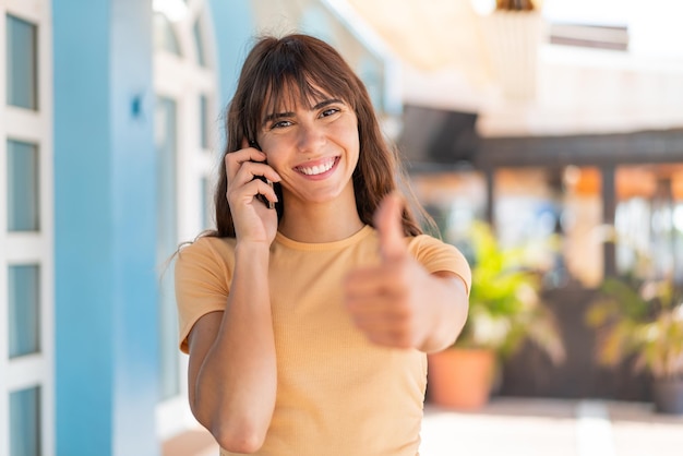 屋外で親指を立てながら携帯電話で会話を続ける若い女性