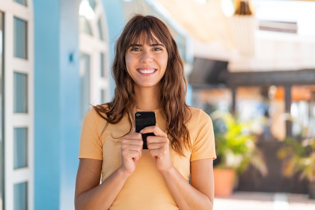 屋外でカメラを見て、携帯電話を使いながら微笑む若い女性