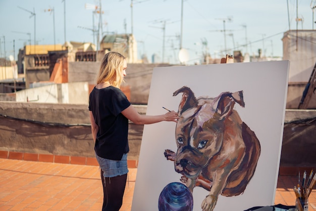 Foto artista della pittura della giovane donna che disegna a casa il tetto. immagine di bulldog su grande tela. arte all'aperto