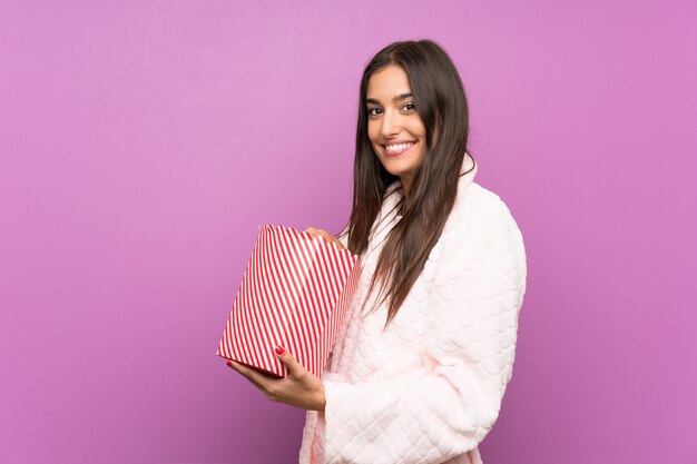 Young woman in pajamas and dressing gown over isolated purple wall holding popcorns