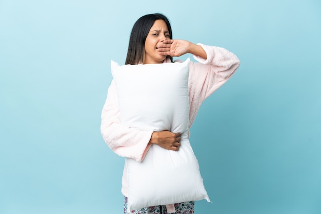 Молодая женщина в пижаме над изолированной стеной в пижаме, держащая подушку и зевая