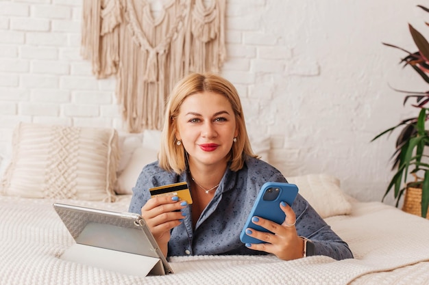 Young woman in pajamas sits on the bed with a computer and a phone makes an online payment