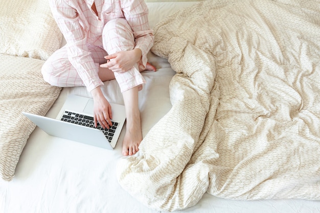 Giovane donna in pigiama che si siede sul letto a casa che lavora usando sul computer pc portatile