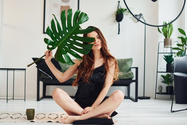 Young woman in pajamas sitting on the floor indoors at daytime and holding big green leaf