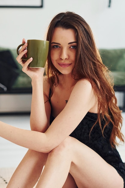 Young woman in pajamas sitting on the floor indoors at daytime with cup of drink