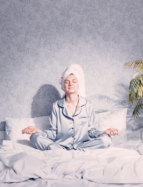 A young woman in pajamas and a towel on her head sits in the lotus position on the bed
