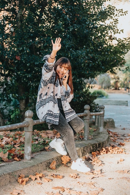 Young woman in the park during a phone call smiling receiving\
good news and saluting to camera on trendy and modern clothes with\
copy space to add your advertisement or copy text.blurred\
background