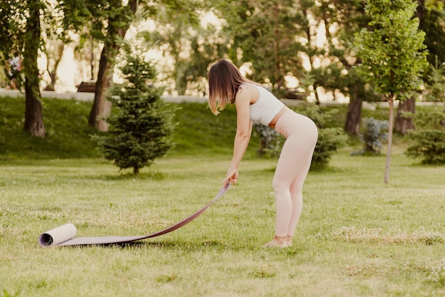 Young woman in park on warm summer morning Woman in sports clothes spreads her yoga and Pilates mat on grass in nature for sports training