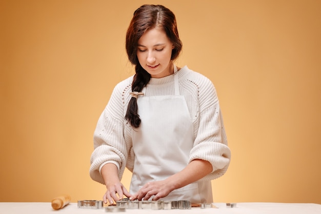 Young woman pastry chef on yellow background cuts christmas cookies in metal shapes