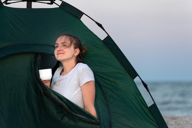 Young woman peeking out of touristic tent girl with cup in the\
tent summer vacation and travel trip