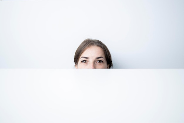 Photo young woman peeks out from under a white cardboard against a white wall