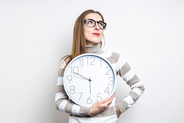 Young woman pensive looking up holding a clock on a light background