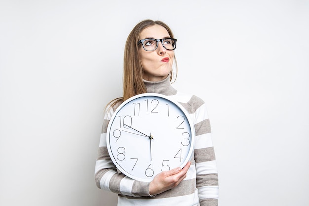 Young woman pensive looking up holding a clock on a light background