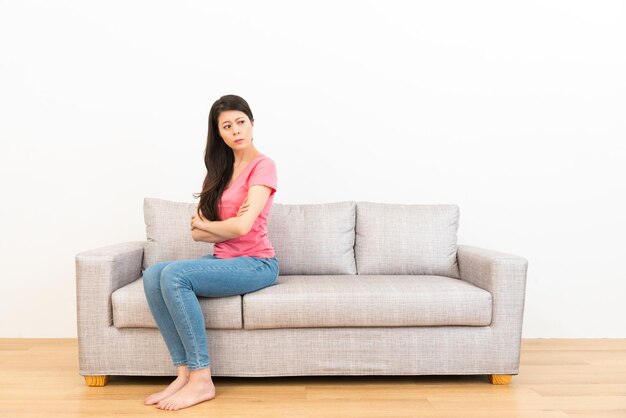 Young woman performance angry emotion sitting on couch sofa\
showing quarrel posture on white wall background with wooden\
floor.