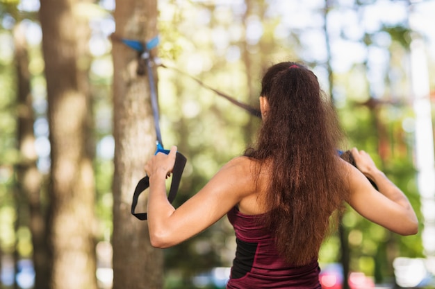젊은 여자는 공원에서 나무에 부착 된 피트니스 스트랩으로 등 근육을 운동을 수행