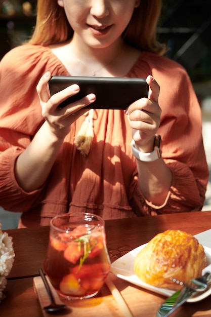 Young woman photographing fruit cocktail and pastry she ordered for lunch in cafe