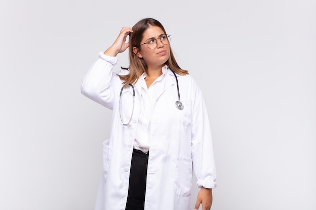 Young woman physician feeling puzzled and confused, scratching head and looking to the side