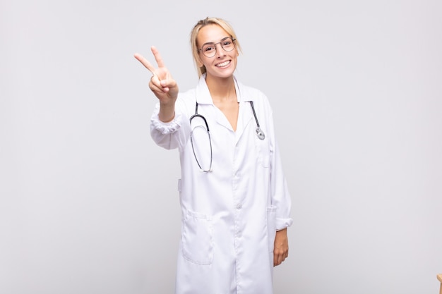 Young woman physician smiling and looking happy, carefree and positive, gesturing victory or peace with one hand